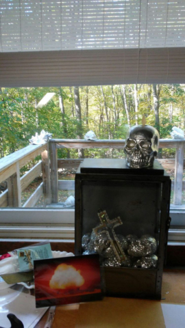 art piece in cabinet with cross and silver skull atop contrasts with woodsy scene outside window